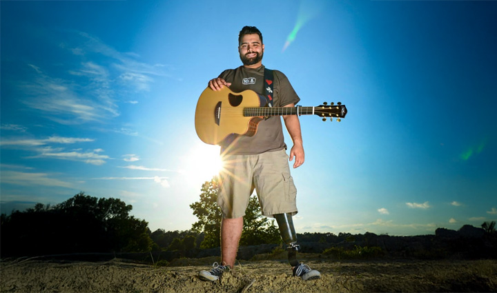 Wounded warrior Sal Gonzalez is backlit by the sun while holding a guitar against the backdrop of a blue sky and palm trees.