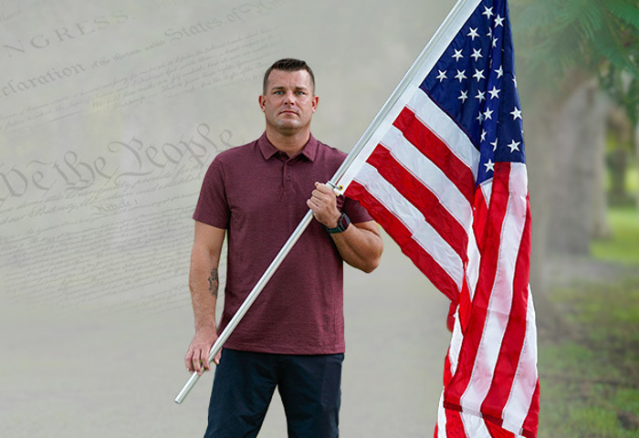 Warrior Mike Larson holds an American flag in a memorial setting with a faded Constitution backdrop and trees.