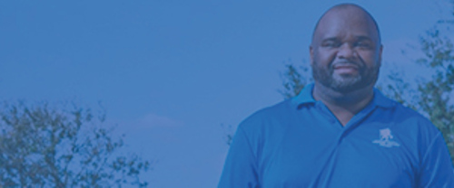 A man dressed in a blue shirt stands against a backdrop of trees, showcasing a peaceful natural setting.