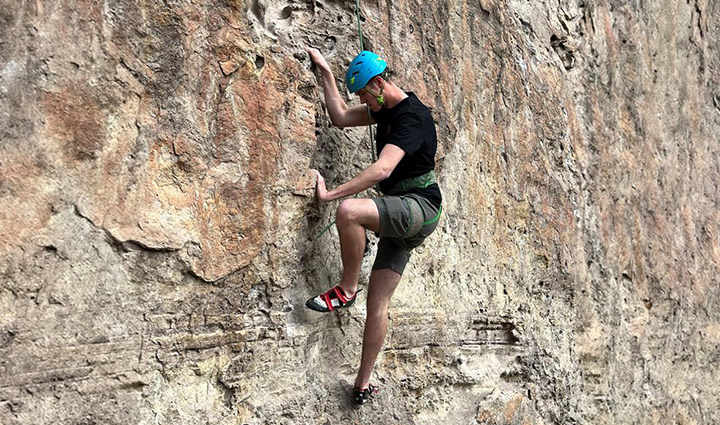 An active-duty service member is rock climbing wearing a blue helmet, black shirt, and gray shorts.