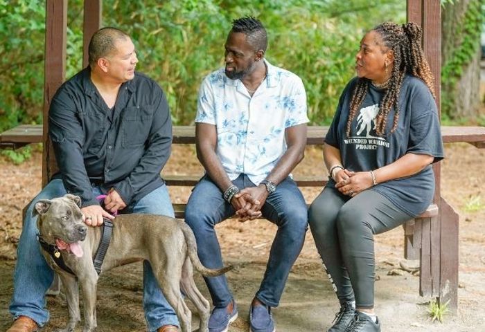 Warriors Eric Frank, Adams Amisu, and Barbara Reese sit outside talking to each other on a bench with Eric's dog at his side.