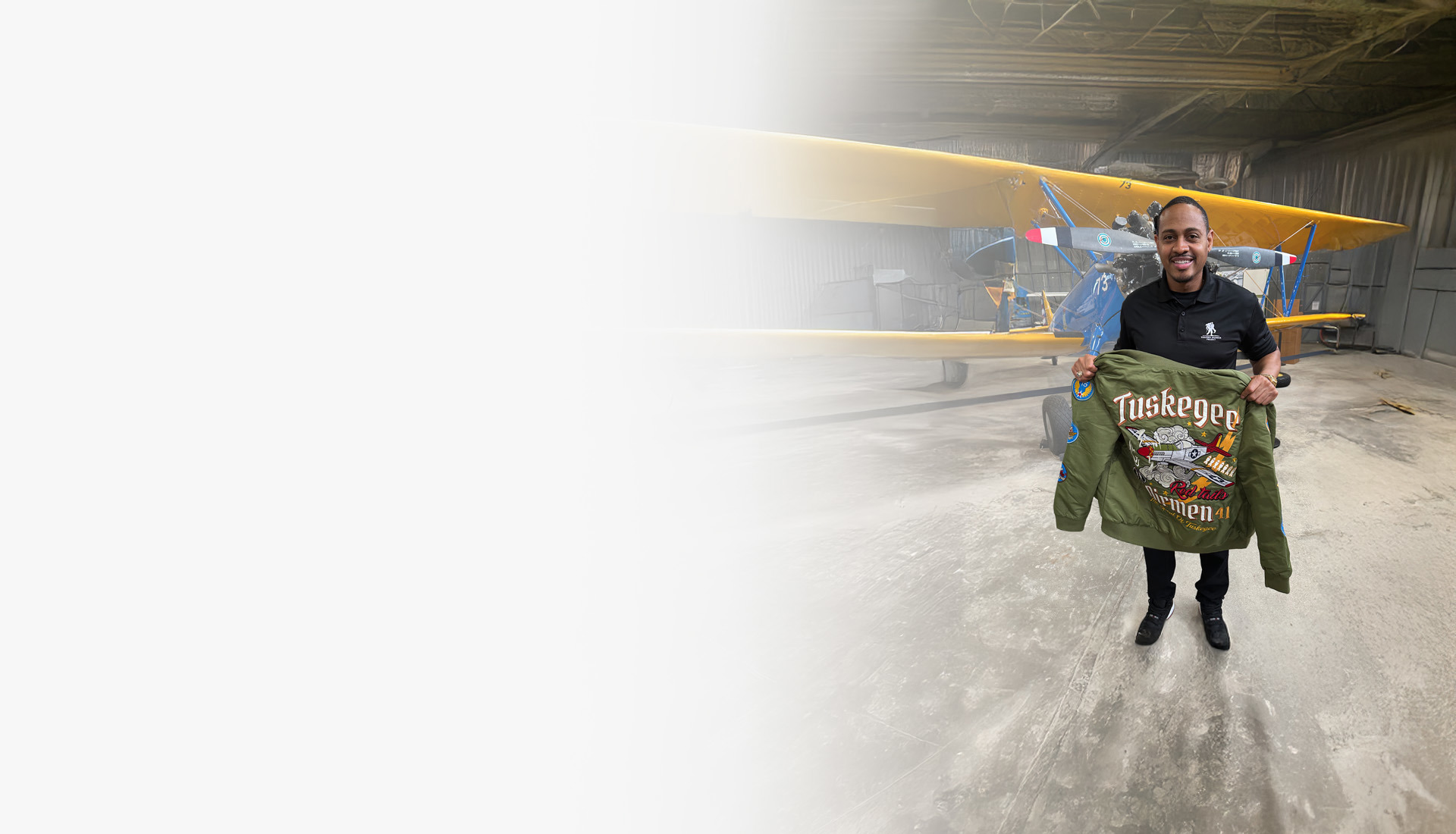 Warrior Tyshawn Jenkins holds a jacket that features the Tuskegee Airmen insignia in front of a blue biplane with yellow wings.