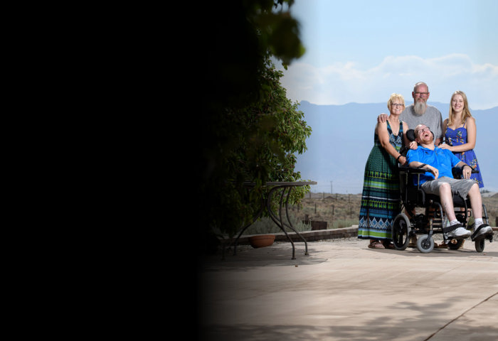 Warrior Erik Schei smiling while in his wheelchair surrounded by his family. They are outside with mountains behind them.