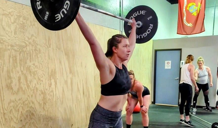 A woman lifts a barbell overhead in a CrossFit gym during the Murph challenge fundraiser, surrounded by other participants.