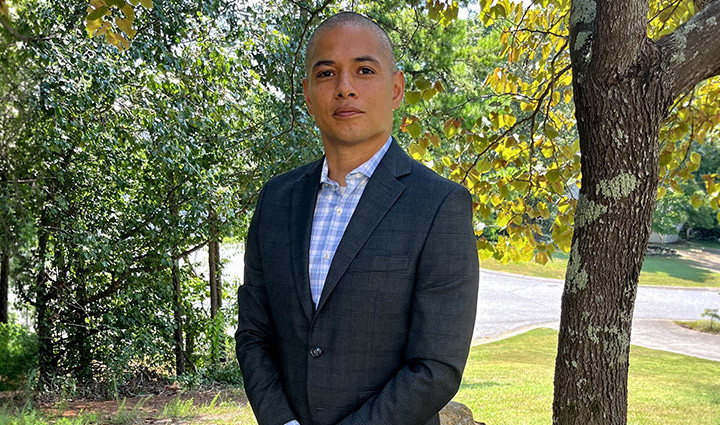 Warrior Daniel Avalos stands next to a tree outside while wearing a dark suit, light collared shirt, and brown dress shoes.