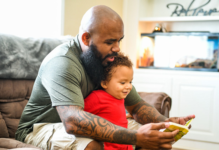 Warrior Eric Delion sits on a couch, wearing a green shirt and khaki shorts while reading to his son, who is wearing a red shirt and khaki shorts.