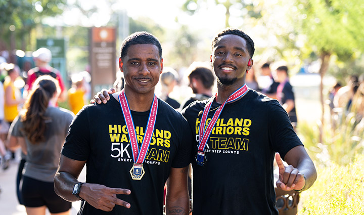 Two male Carry Forward 5K participants wearing ‘Highland Warriors 5K Team – Every Step Counts’ shirts pose with their medals.