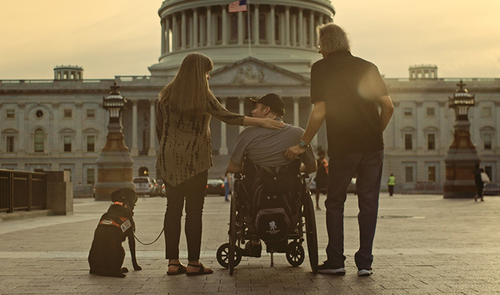 Veteran using a wheelchair with two caregivers and a service dog in front of the U.S. Capitol at sunset. 
