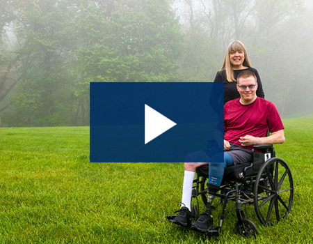Wounded warrior Jason Ehrhart sits in a wheelchair while his mother and caregiver Pam Estes stands behind him smiling.