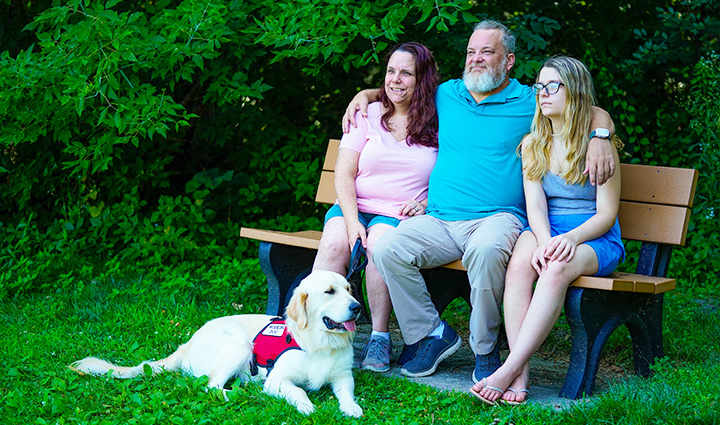 Warrior David Denhardt sits on a bench with his wife and daughter, smiling, with his service dog sitting on the ground.