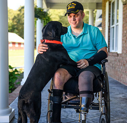 Seated in a wheelchair, wounded warrior Jason Ehrhart smiles as he gazes downward at his black labrador.