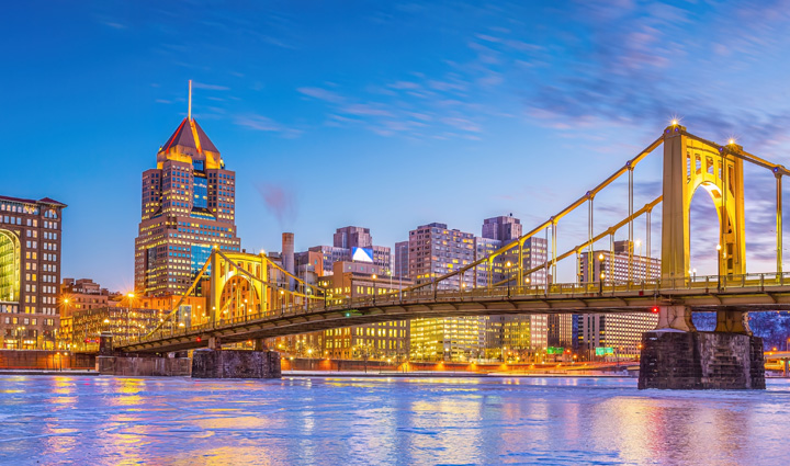Illuminated yellow suspension bridge spanning a partially frozen river at dusk, with the Pittsburgh, Pennsylvania skyline glowing in the background.
