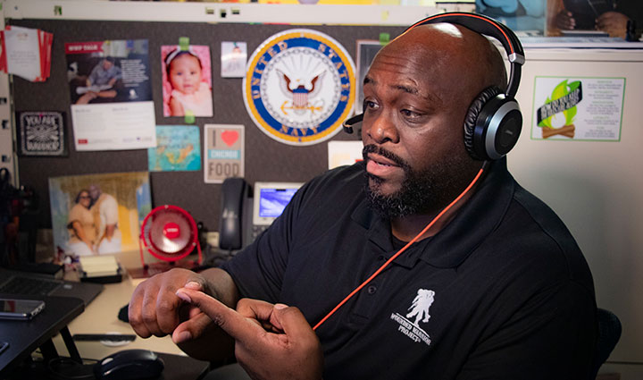 A member of the WWP Mental Health team speaks during a virtual call, wearing a headset and Wounded Warrior Project polo. Seated at his decorated cubicle, he gestures with his hands while engaging in conversation.