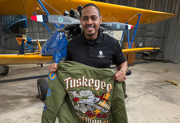 Warrior Tyshawn Jenkins holds a jacket that features the Tuskegee Airmen insignia in front of a blue biplane with yellow wings.