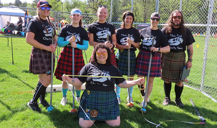A group of seven individuals, dressed in kilts and black t-shirts, pose with heavy implements for the Pike's Peak Charity Games on a grassy field.