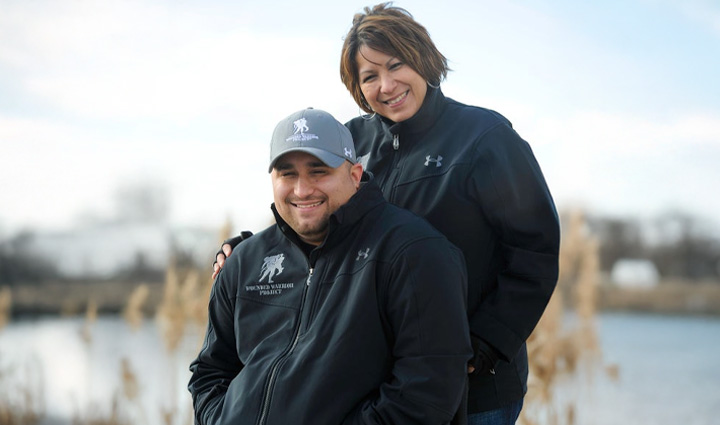 Caregiver Cindy Parsons stands behind her son, Shane, with her arm around him. Shane sits on a wheelchair and wears a black WWP jacket and a gray WWP cap.