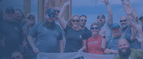 Group of people outdoors posing together behind a banner, some with sunglasses, smiling and raising their hands.