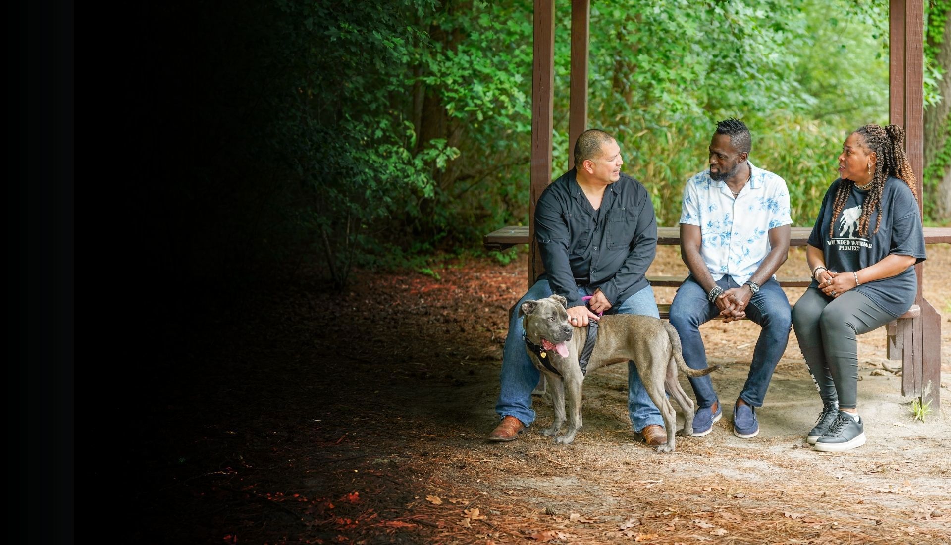 Warriors Eric Frank, Adams Amisu, and Barbara Reese sit outside talking to each other on a bench with Eric's dog at his side.
