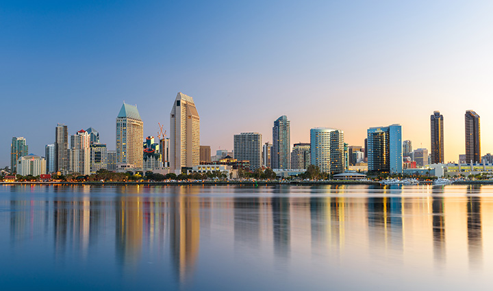 San Diego skyline at sunrise or sunset, with high-rises reflected in calm water under a clear sky with blue-orange gradient.