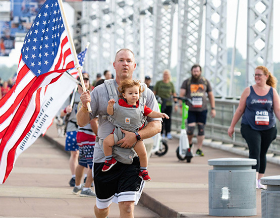 A man carries a flag and a baby in a chest carrier while running a Carry Forward 5K across a bridge.