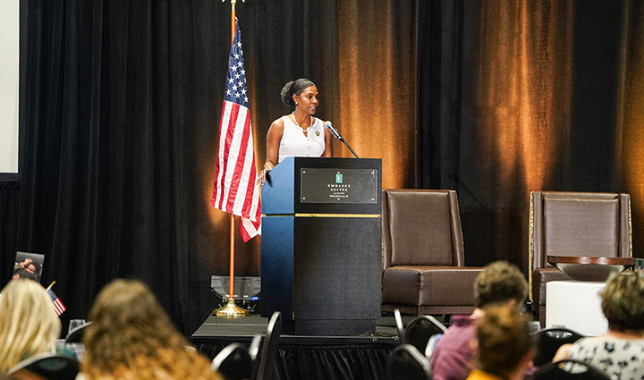 Wounded warrior Tonya Oxendine, dressed in business attire, standing next to an American flag, speaks on stage at the Battlemind to Home summit in Indiana.