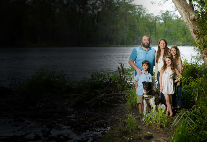 Family support member and caregiver, Jenna Malone, her husband Issac and their children stand next to tree smiling at camera.