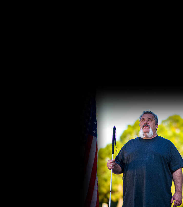 Wounded warrior Aaron Cornelius standing outside holding a cane beside an American flag