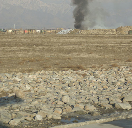 Dry, barren landscape with scattered rocks in the foreground. Background shows smoke rising from a distant mound, with mountains under a hazy sky.