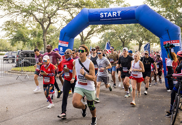 A group of runners begin a Carry Forward 5K event under a blue ‘Start’ arch.