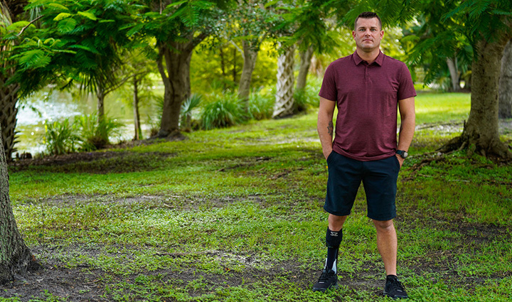 Warrior Mike Larson is standing in a wooded background, with his hands in his pockets, wearing a maroon shirt, black pants, and black shoes.