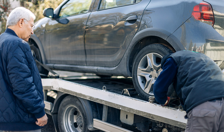 A gray car is loaded onto a tow truck as the driver secures it and the owner watches attentively.