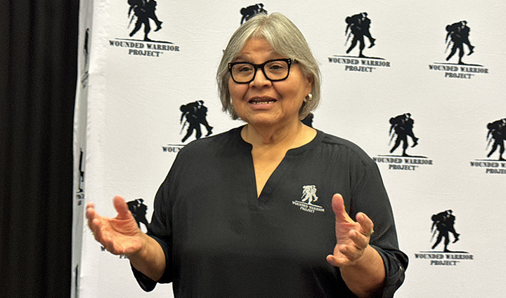 Caregiver Mary Tallouzi shares her story on stage at an event. She wears a black WWP blouse and black-rimmed glasses.