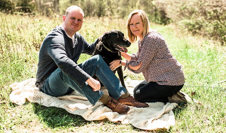 Caregiver Jennifer Jenkins is seated on a picnic blanket alongside her brother, James Smith, with James’ black lab service dog between them. 