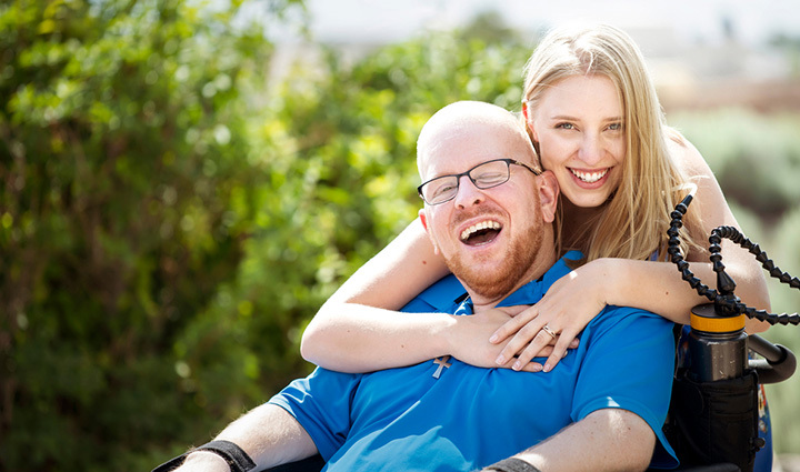 Warrior Erik Schei sitting in a wheelchair and smiling while his sister hugs him.