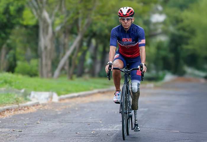Warrior Kelly rides a road bike on a tree-lined street, wearing a USA cycling kit and helmet, with a prosthetic leg.