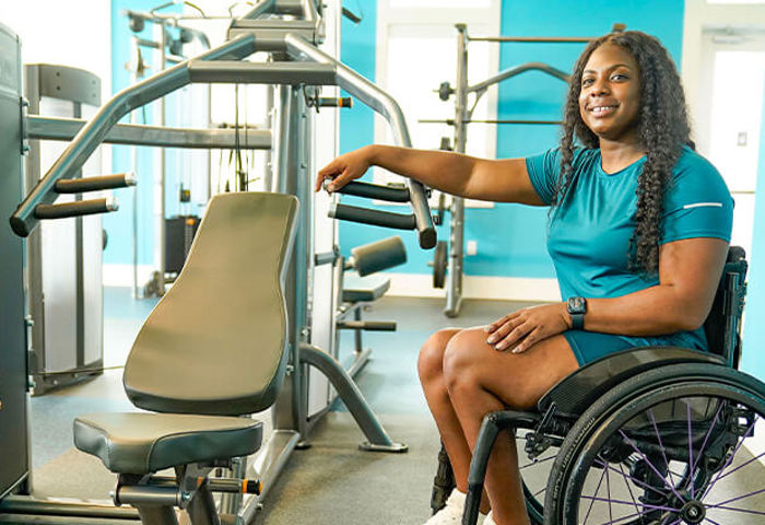 Warrior Corine Hamilton sits in a wheelchair in a gym, smiling at the camera while draping her arm over the a shoulder press machine.