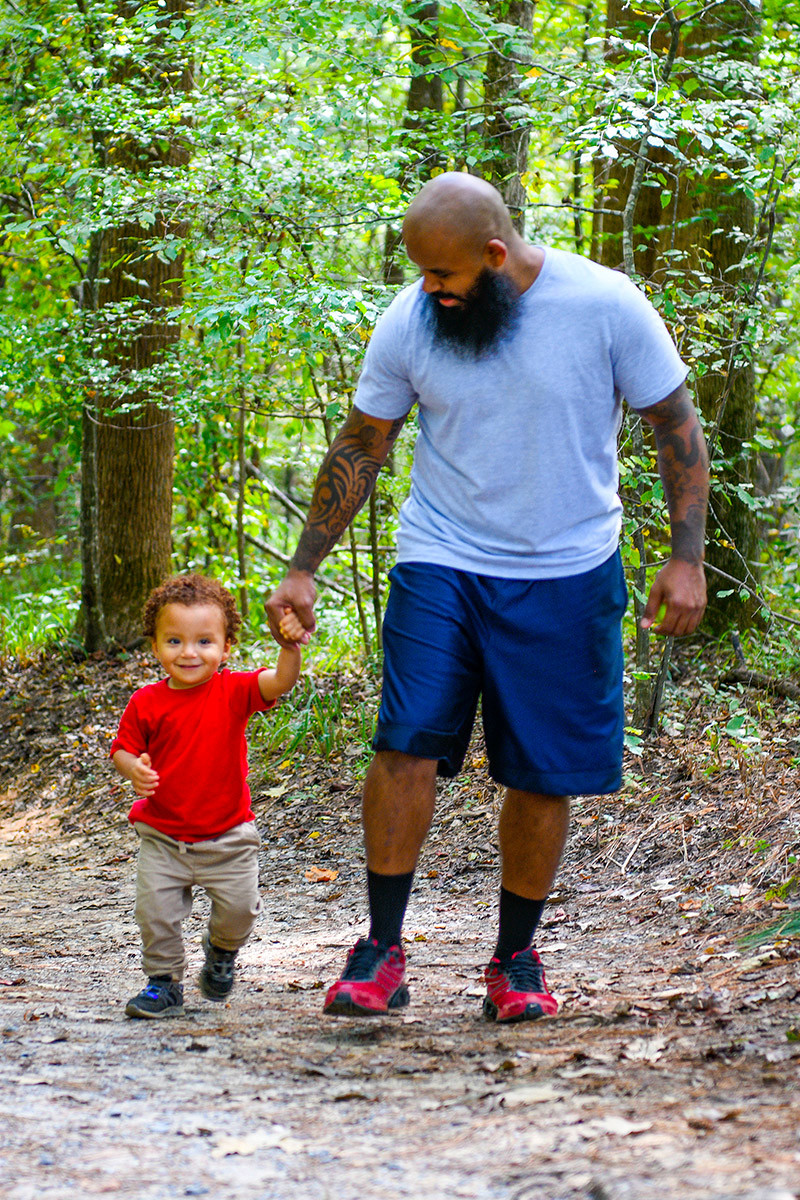 Wounded warrior Eric Delion holds his toddler’s hand while walking with him along a path surrounded by trees. He smiles down at his son as they walk.