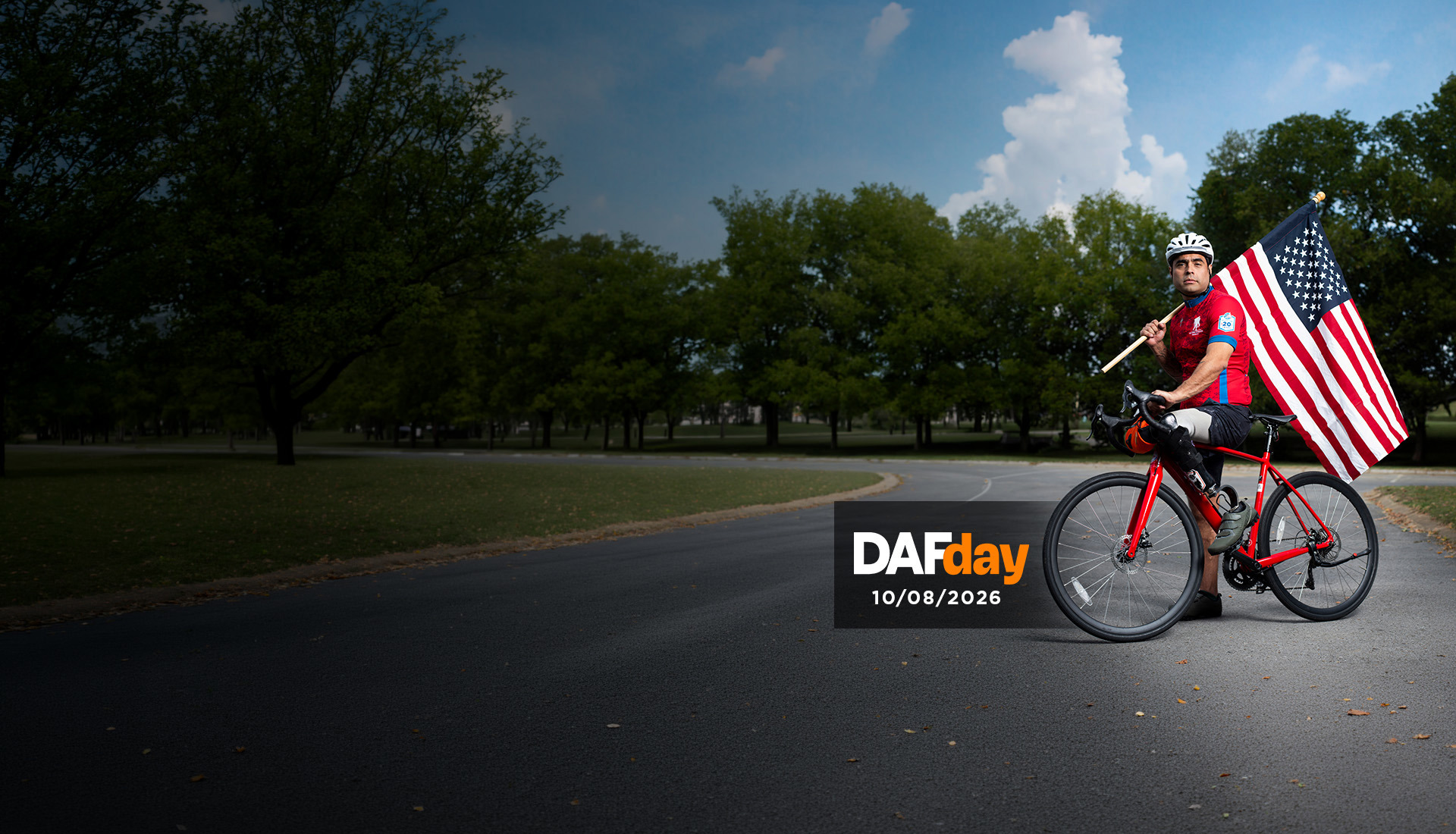 Warrior, Sal Gonzalez on a red bike with his prosthetic leg, holding a large American flag on a paved road with trees in the background.