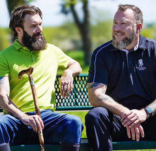 Two men with beards sit on a bench outdoors. One wears a green shirt and holds a cane; the other wears a dark polo. They share a relaxed conversation.