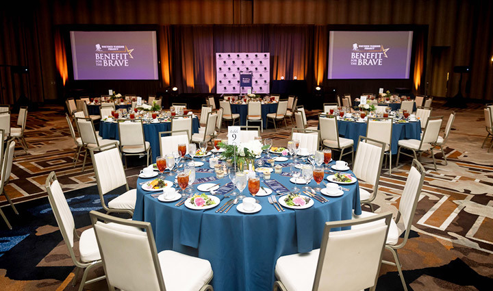 Round banquet tables set for a formal event fill a ballroom, each covered with blue tablecloths, white chairs, and neatly arranged place settings with plates, glassware, and iced tea. A stage with podium and two large screens displays ‘Benefit for the Brave’ against a backdrop of warm orange lighting.