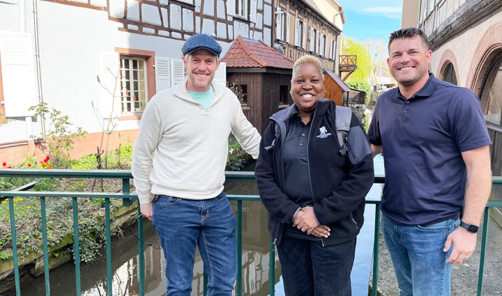 Warriors John Rego, Donna Pratt, and Mike Larson stand in front of a river in between two buildings while smiling.