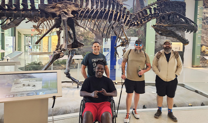 Four veterans smile in front of a dinosaur exhibit at a local San Antonio museum, including one using a wheelchair.