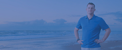 A person stands on a beach near the shoreline, wearing a blue polo shirt and a watch, with the ocean and clouds in the background.