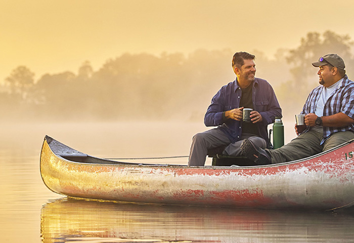 Warrior Tim Aponte and a fellow veteran sit at the edge of a dock at sunrise with their feet resting in a canoe.