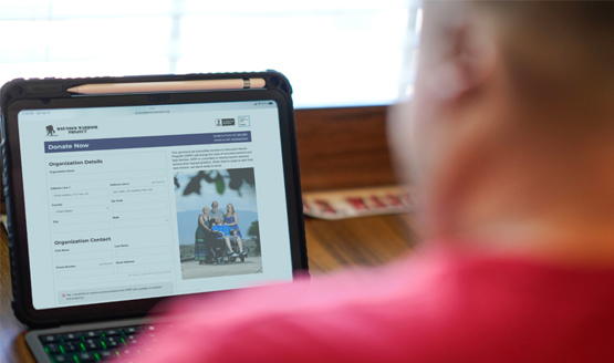 Man with red shirt sits at a table with a tablet open to complete an online donation form to Wounded Warrior Project. The screen displays fields for organization and contact details.