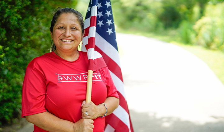 Wounded warrior Maria Edwards wears a red WWP Fit t-shirt while smiling and holding an American Flag along a tree-lined path.