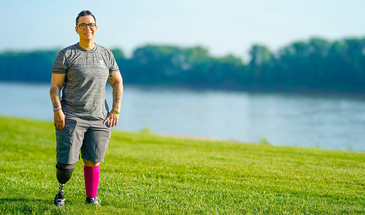 Female warrior Beth King stands before a lake, her prosthetic leg visible as she faces the camera with strength and confidence.